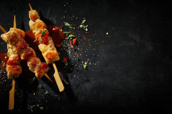 Three healthy chicken breast kebabs with chili sauce seasoned with spices and fresh chopped herbs on wooden skewers viewed close up in square format on dark grey